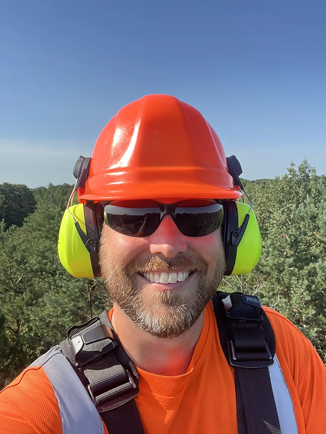 Shawn Cagle above tree tops in a boom lift with helmet, eye, and ear protection.